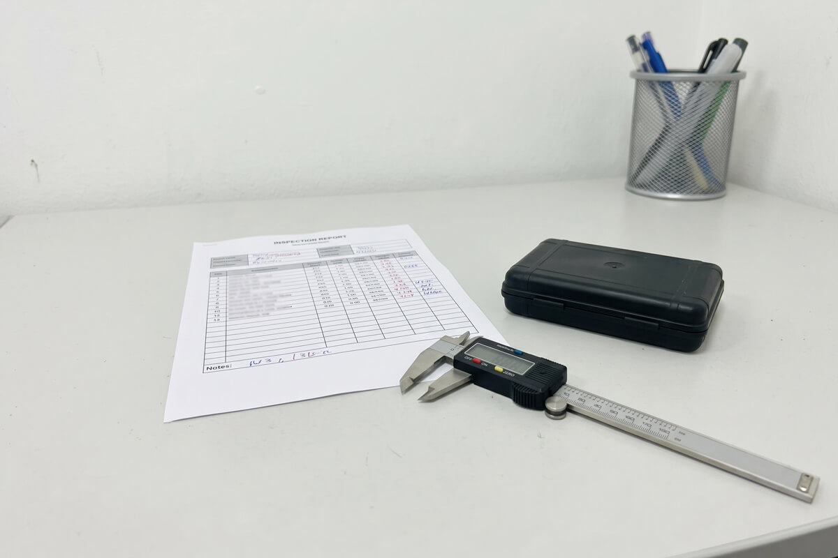Printed first-article inspection report beside a black molded sample part and a digital caliper on a desk