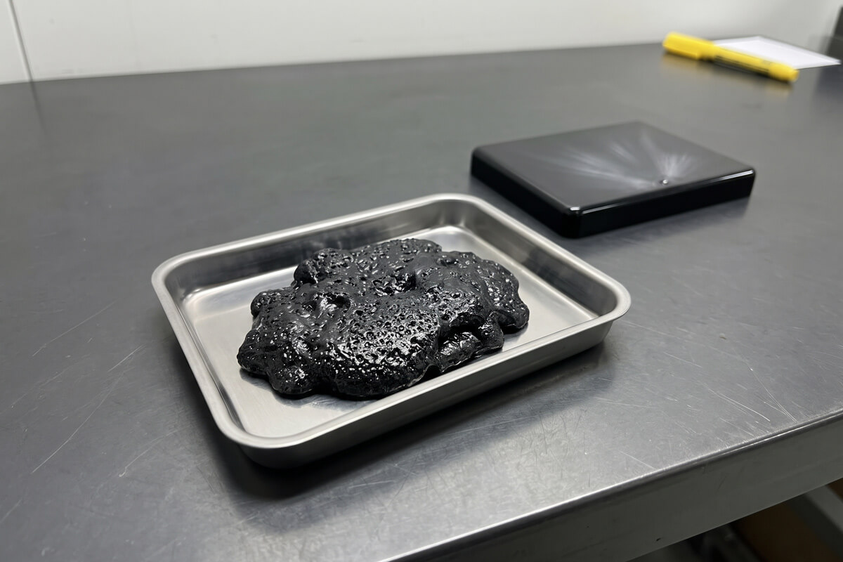 A black purge lump full of small bubbles on a stainless steel tray beside a glossy molded lid