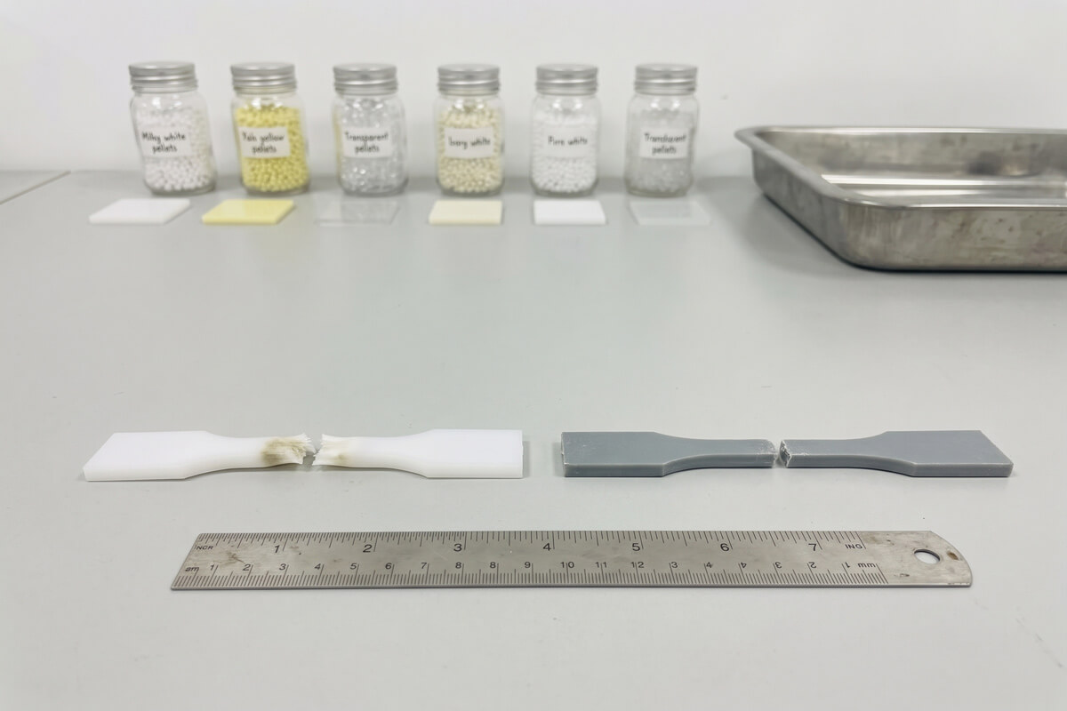 Two broken tensile test bars, one unfilled white and one glass-fiber-filled gray, on a lab bench beside a ruler