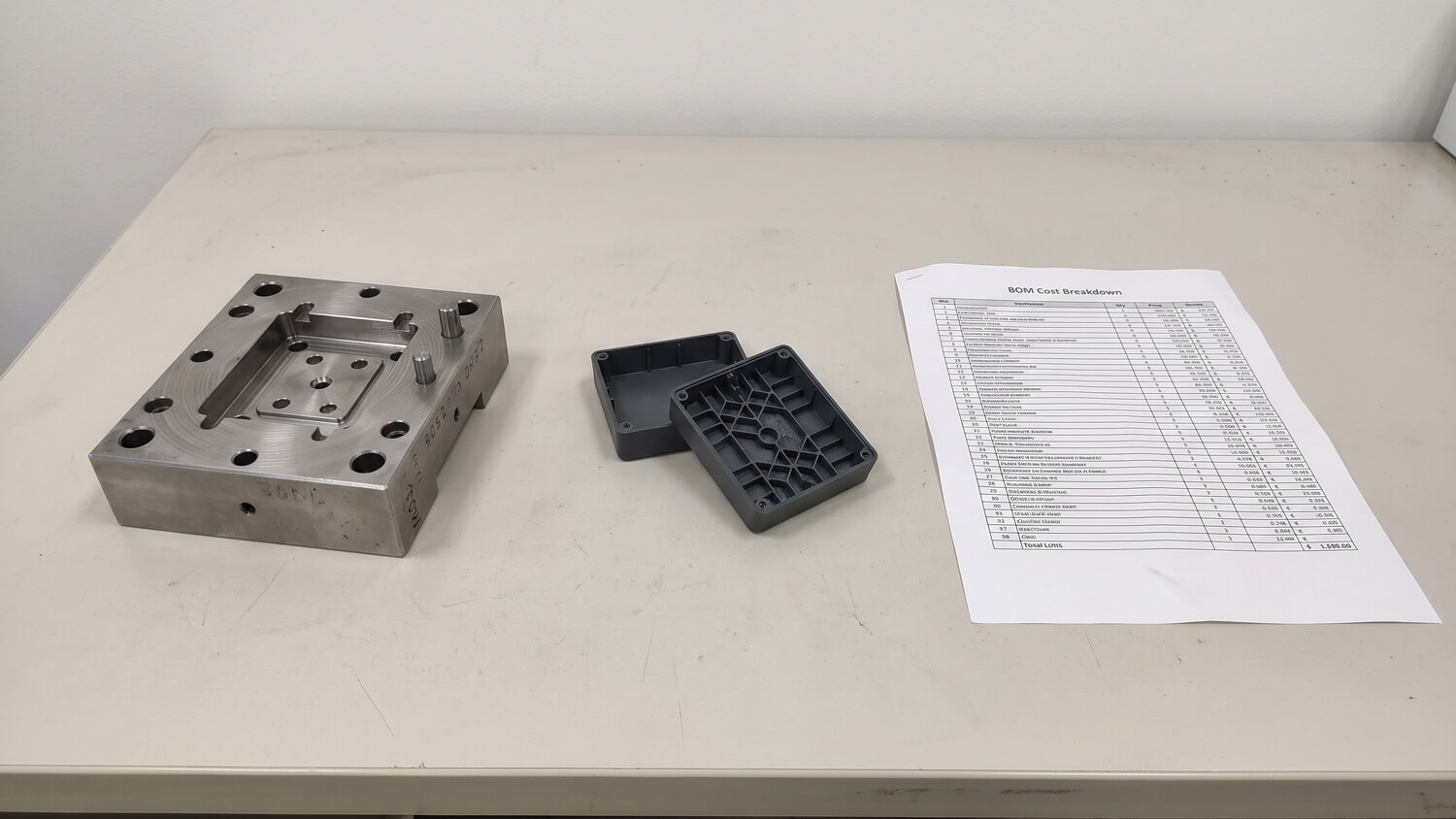 MUD rapid tooling steel insert alongside injection-molded plastic accessory housings and a BOM cost analysis sheet on an office desk