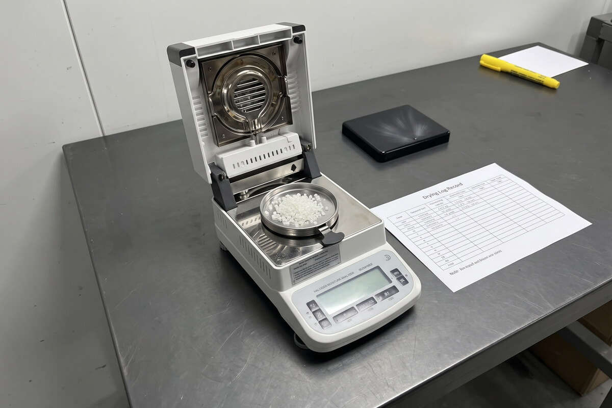 A benchtop moisture analyzer with a small pellet sample dish beside a printed dryer log sheet