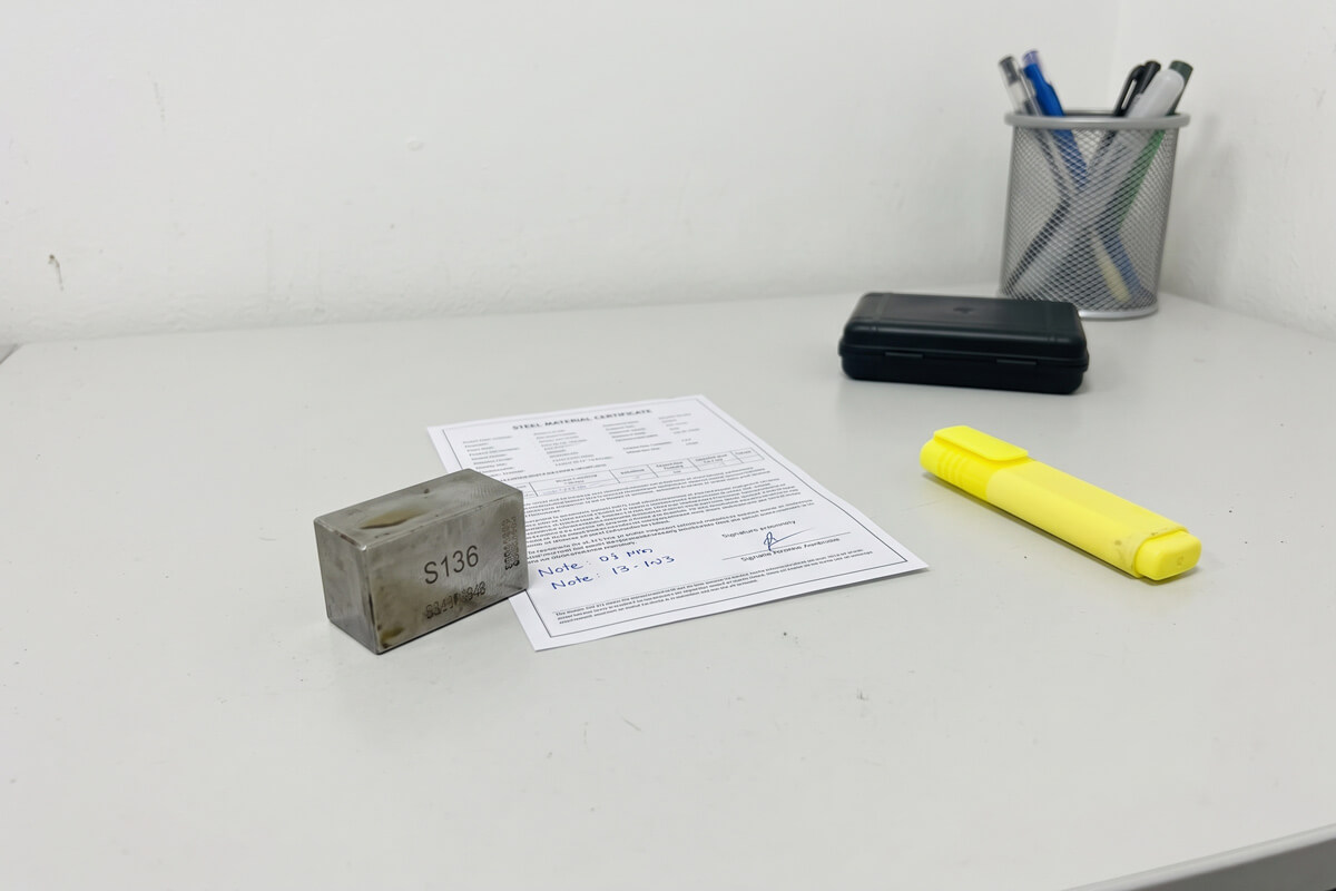 Small mold steel insert with stamped grade marking beside a printed steel mill certificate on a desk