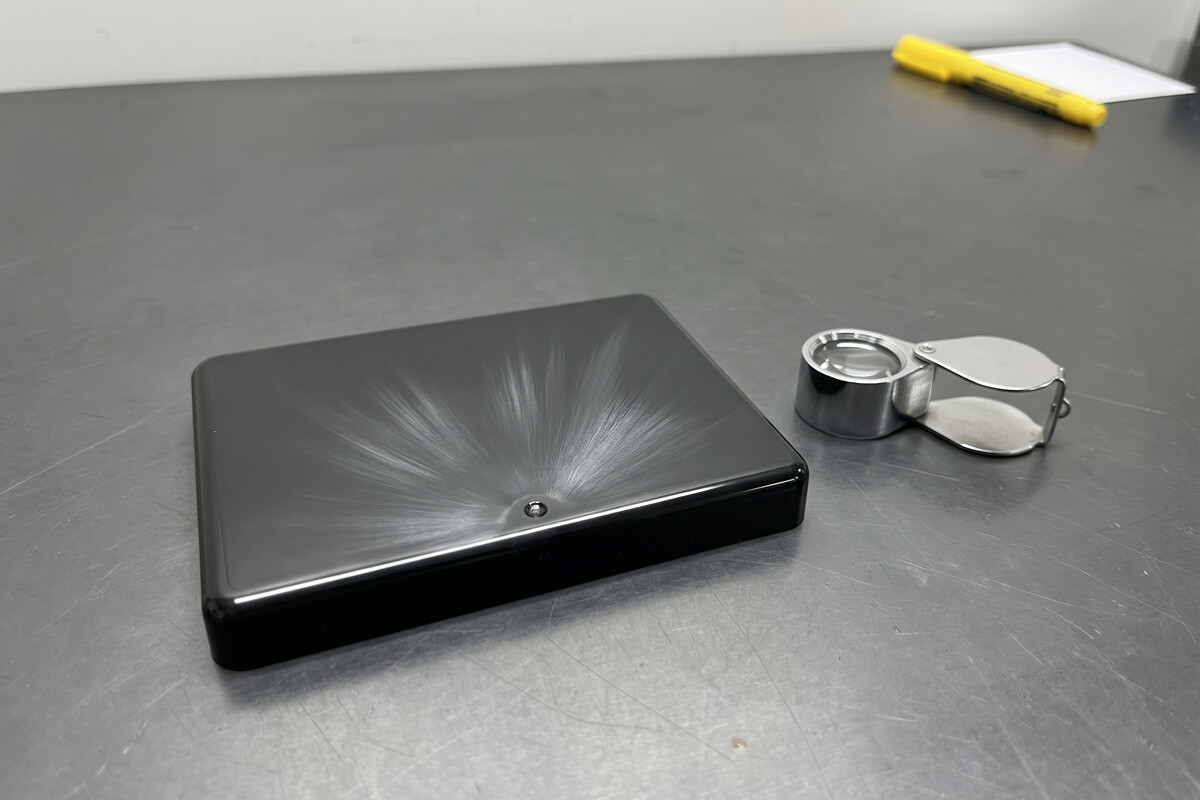 Close-up of silver splay streaks radiating from the gate area of a glossy black molded lid, with a loupe beside it