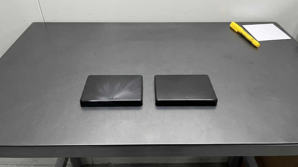 Two glossy black injection molded lid parts side by side, one with silver splay streaks near the gate and one clean
